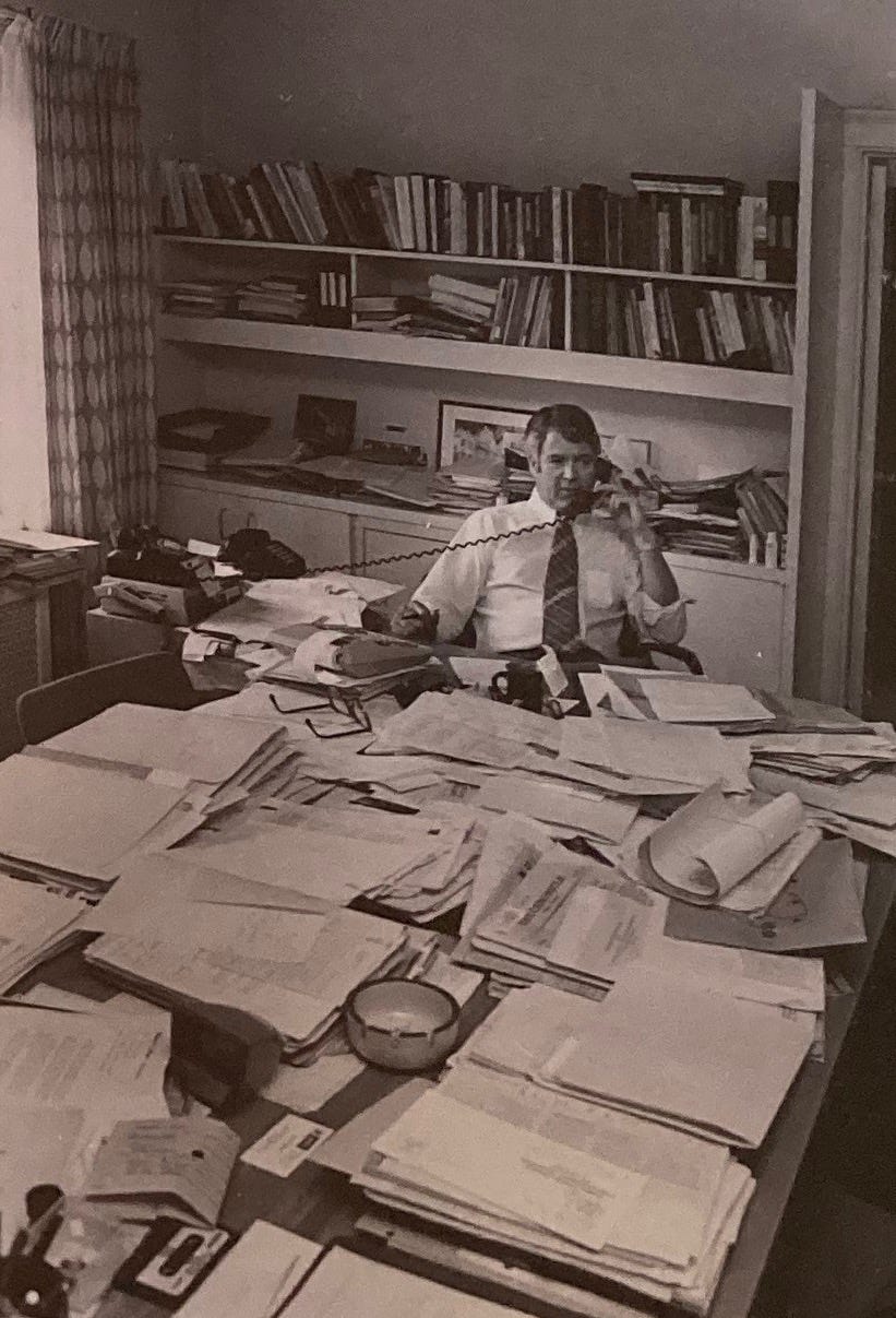 Siloh's grandfather as a middle-aged man at a desk covered with papers, and talking on the phone