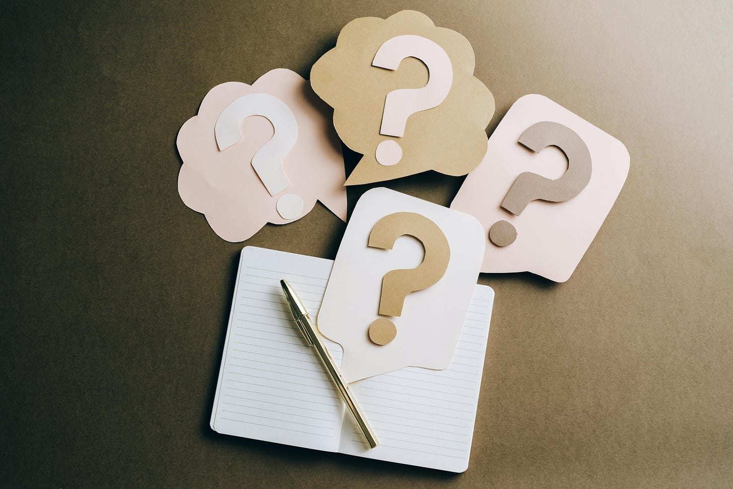 An open notebook, with a gold pen inside it, there are four question marks floating on construction paper coming out of the notebook. The pallet is neutral, calm, and earthy. 