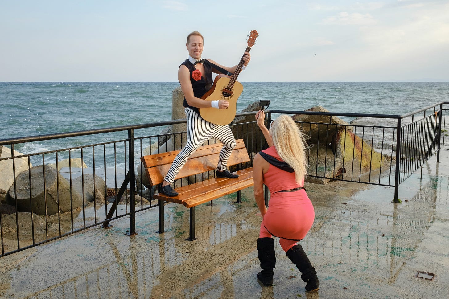 A man in odd black and white lined trousers stands on a bench holding a guitar while a woman films him.