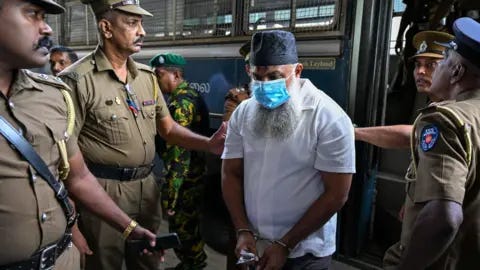 AFP via Getty Images A man with a beard, skullcap and face mask being led out of a bus by four police officers