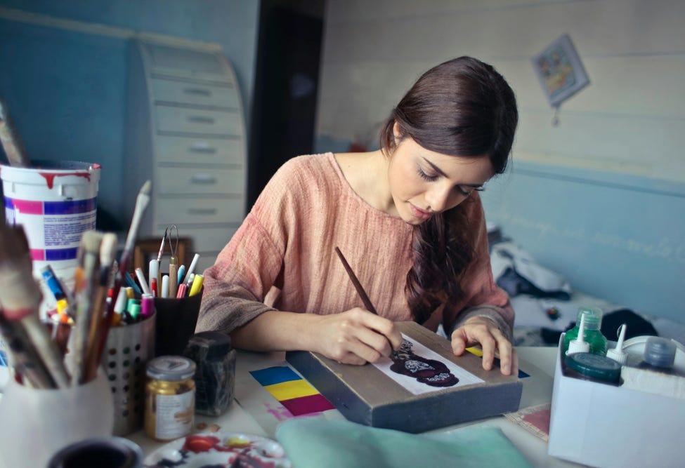 A woman with long dark hair wearing a peach shirt painting on a canvas at her desk.