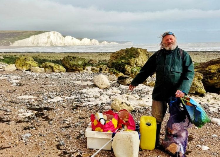 Man wearing dark jacket on a pebbly beach, sorting and collecting trash into boxes and bags with white cliffs in the background.