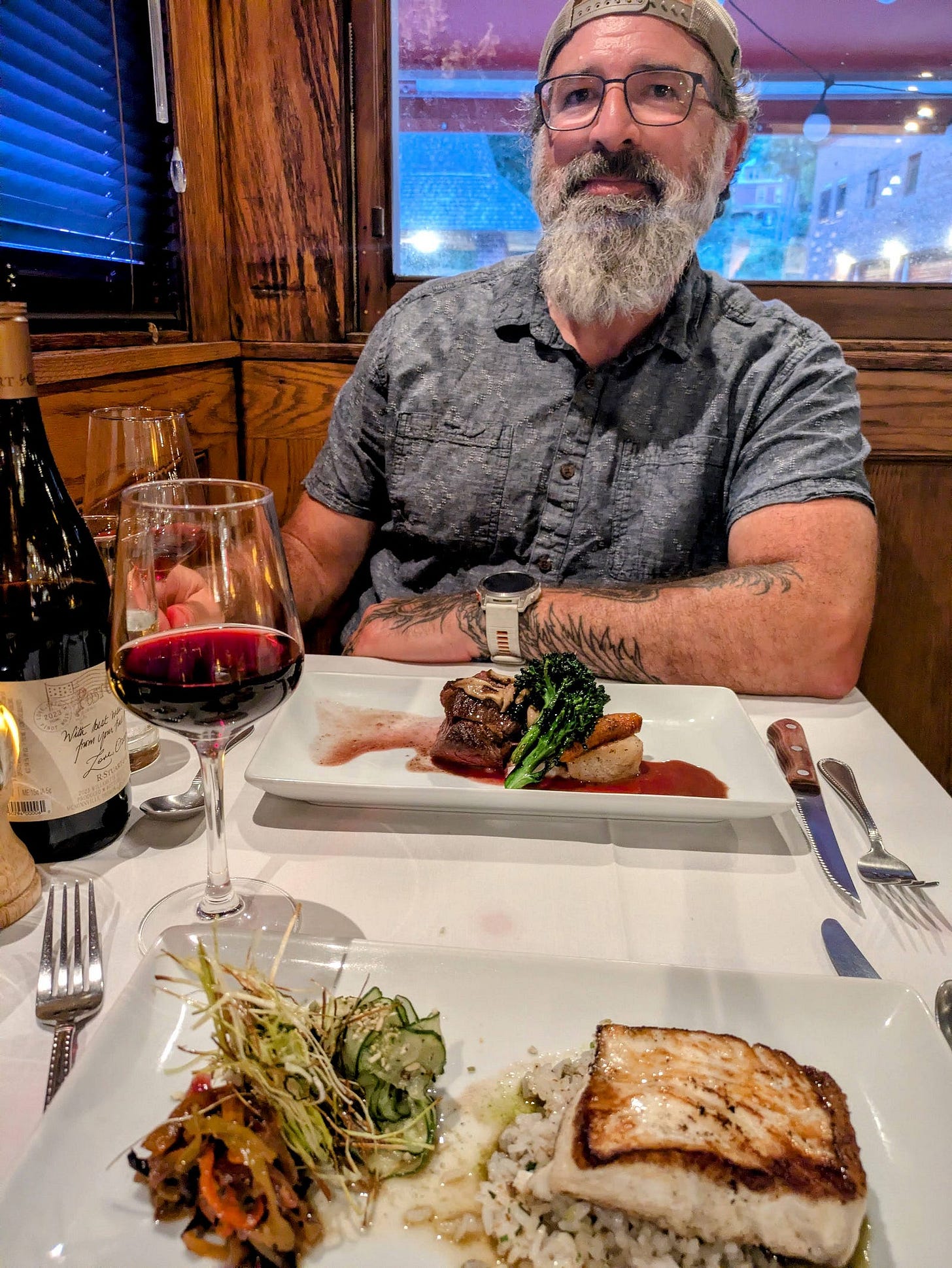 The author smiles at a candlelit dinner table inside T.J. Buckley’s restored 1925 dining car, wearing glasses, a gray shirt, and a tan cap, his gray beard prominent. Two elegantly plated entrées—seared meat with broccolini and pan-seared fish over rice—sit on the white tablecloth alongside a bottle and glass of red wine. The author smiles at a candlelit dinner table inside T.J. Buckley’s restored 1925 dining car, wearing glasses, a gray shirt, and a tan cap, his gray beard prominent. Two elegantly plated entrées—seared meat with broccolini and pan-seared fish over rice—sit on the white tablecloth alongside a bottle and glass of red wine.