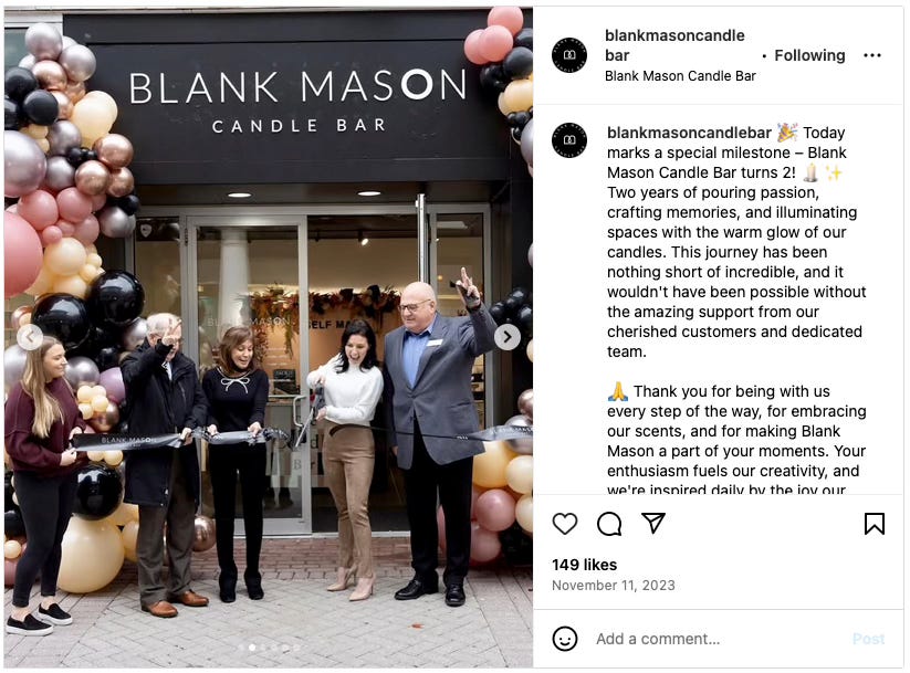 Instagram post from blankmasoncandle showing a ribbon-cutting ceremony at their grand opening. Several people including what appears to be city officials and the owner stand in front of the store entrance decorated with black, pink, and gold balloons. The caption celebrates Blank Mason Candle Bar turning 2 years old.