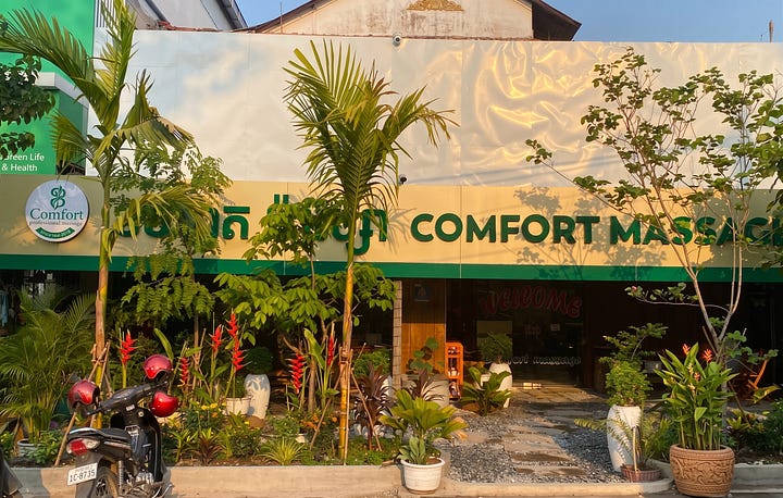 A composite image with a text header that reads, "🧘 Wellness Recommendation: Healing at Comfort Massage." Below the text are two photographs side-by-side. The left photograph shows the exterior of the massage parlor, which has a green facade and a sign that reads "COMFORT M...". It's surrounded by a lush garden with palm trees and shrubs. The right photograph shows the interior foot massage area with a row of blue reclining chairs lined up along a wood-slat wall. Each chair has a footrest with towels, plumeria flowers, and a cluster of stones.