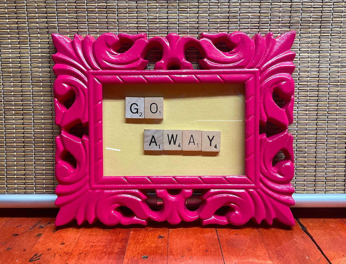 Ornate pink picture frame with scrabble tiles spelling out the words "Go Away" on a yellow background.