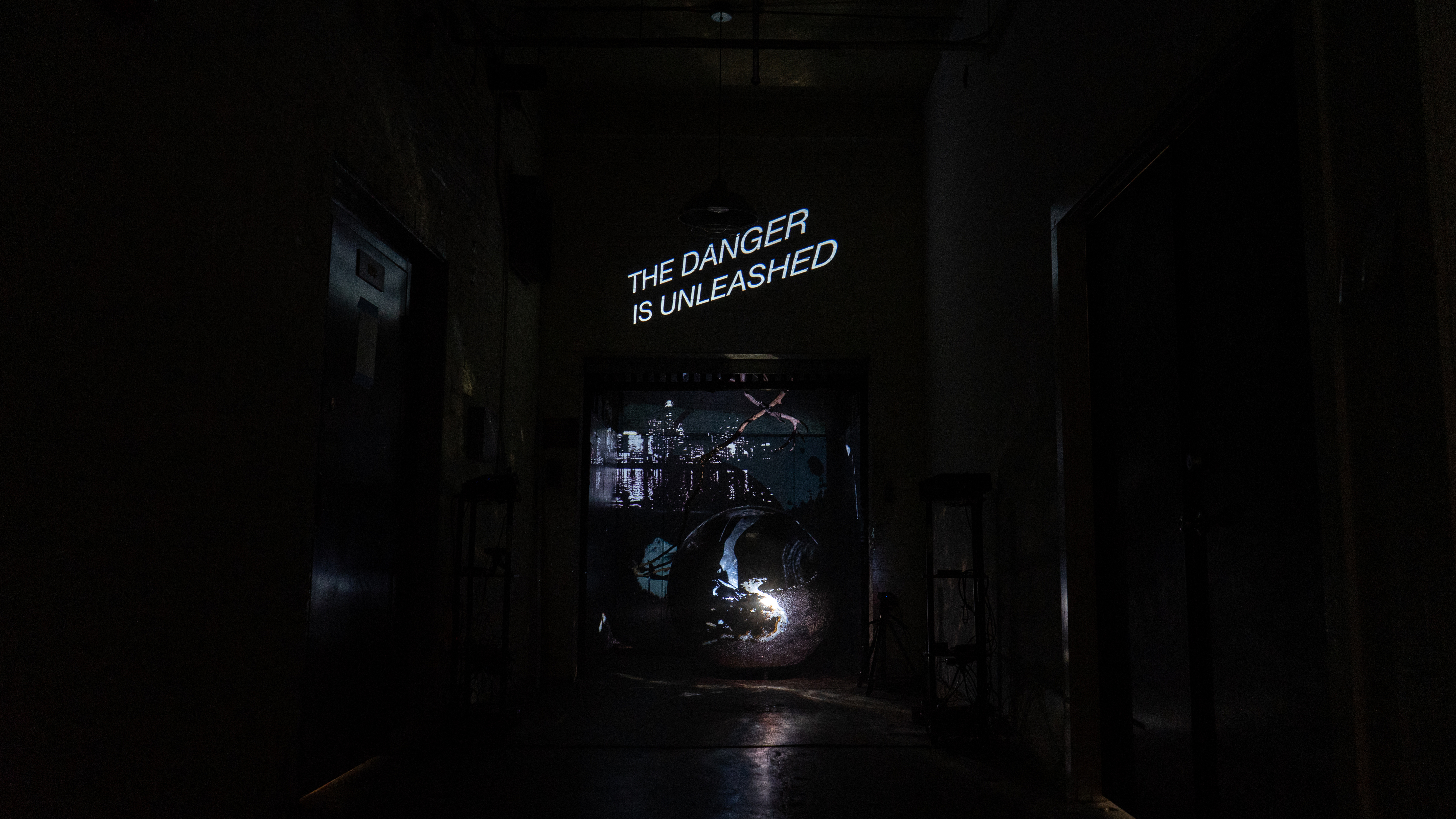 A dark hall leads to a central square opening, the freight elevator. The elevator is transformed into a gallery. Within it is a large metallic sphere lit by overlapping projections depicting water, flame, a city skyline - all in black and white. Above the steel ball, in front of the skyline, a creature is hovering. It is made of wood and has wings that look like antlers. Above the entry to the elevator, on the wall above, white text states, “the danger is unleashed.”