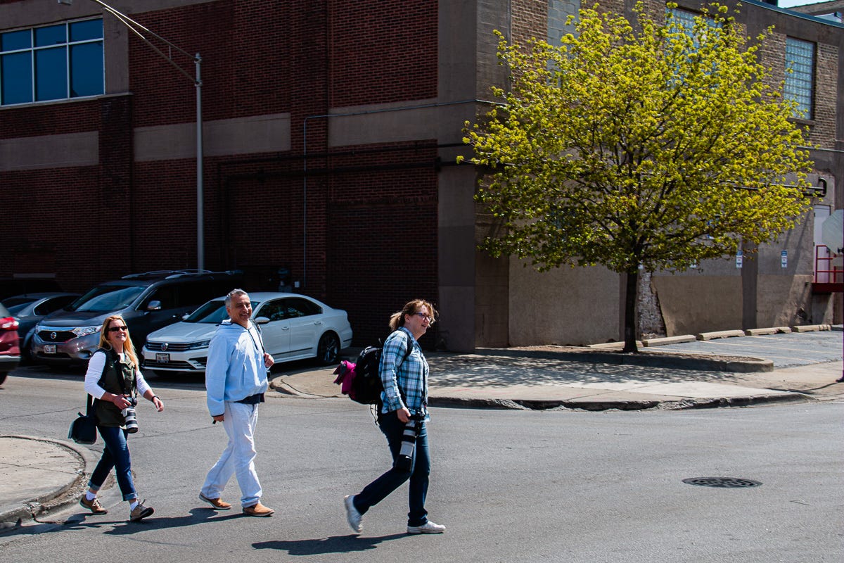 Three people, two women and one man, crossing a street near industrial buildings, carrying cameras and backpacks.