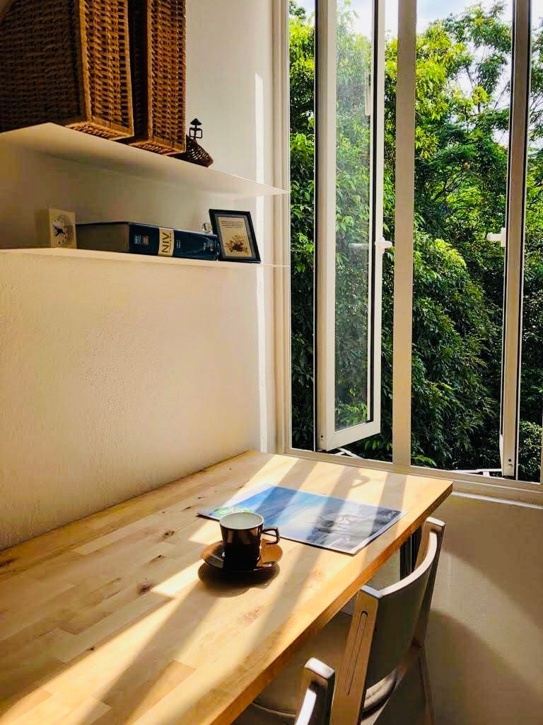 Image: photo of a long, oak bar table, with a side window and trees outside, a glass of coffee on the table