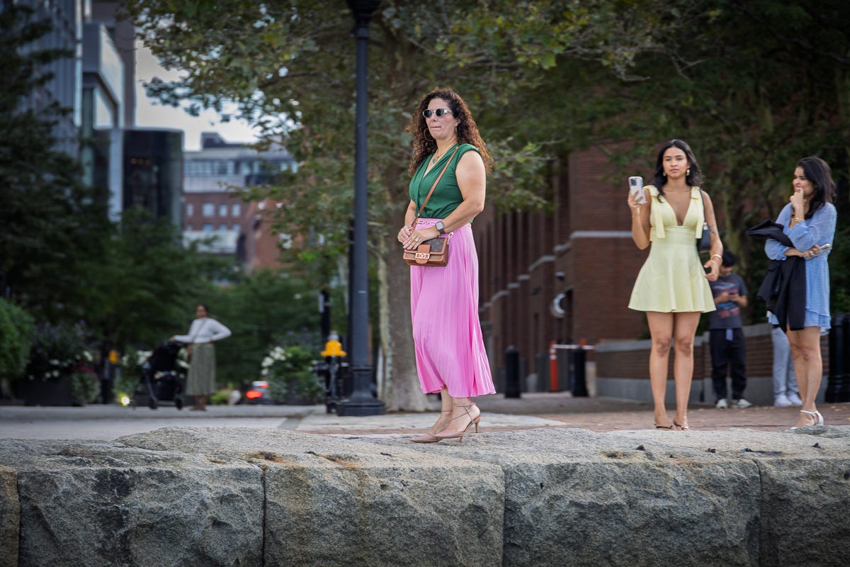 Woman in green top and pink pants standing on Seaport Harborwalk as others watch and take photos nearby