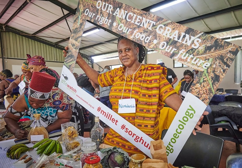 A woman selling vegetables and grains holds up a sign saying 'ancient grains: our right, our food, our future' A woman selling vegetables and grains holds up a sign saying 'ancient grains: our right, our food, our future'