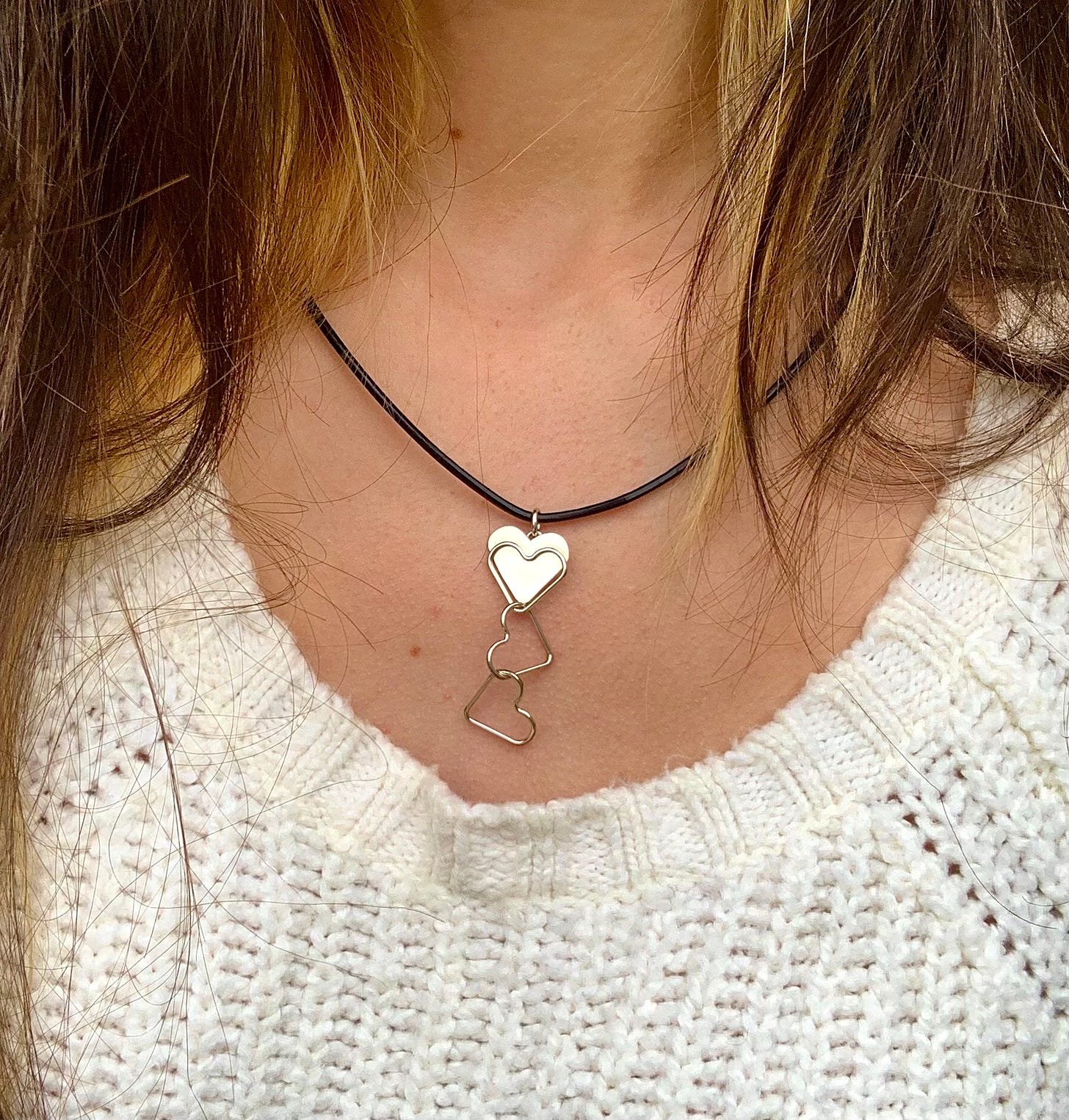 Photo of a silver necklace of three linked hearts, worn by a brown-haired woman in a white sweater