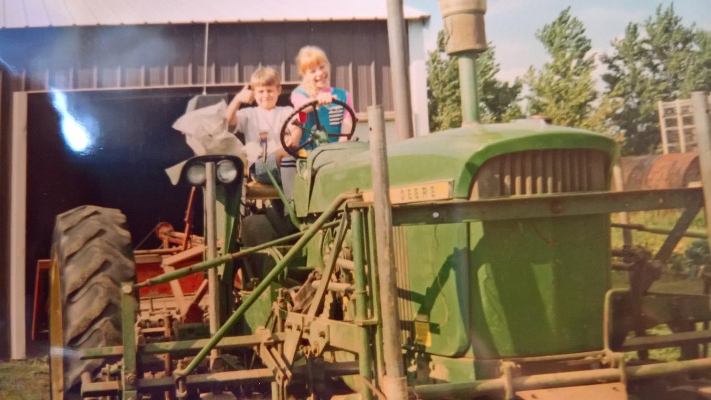 Two kids sit on an old green tractor, smiling and giving a thumbs up.