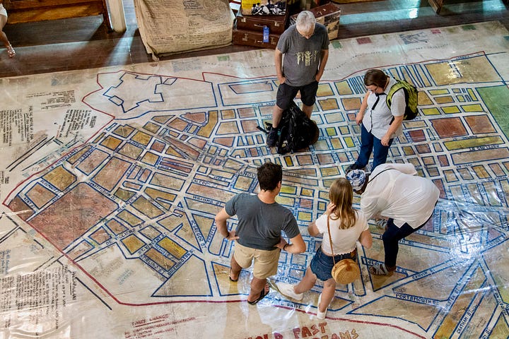 Four photographs from South Africa. Visitors stand on the hand-drawn floor map of old District Six inside the District Six Museum in Cape Town. A steep street of brightly painted houses in Bo-Kaap, with two people walking uphill carrying shopping. A Rovos Rail train alongside a platform at Capital Park station in Pretoria, with green and white branded umbrellas in the foreground. The interior of Clarke's Bookshop in Cape Town, seen from an upstairs landing, with crowded shelves and customers browsing tables of books.