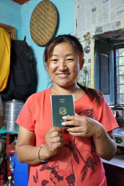 Close up of Nepali woman wearing a red t-shirt and smiling. She is holding up her passport.