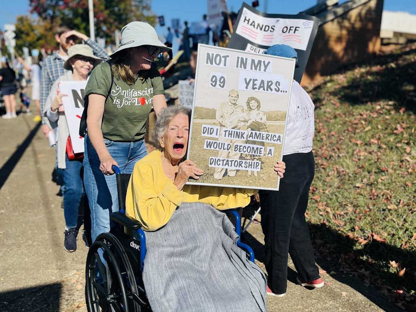 A 99-year-old woman using a wheelchair holds a sign that reads, "Not in my 99 years did I think America would become a dictatorship"