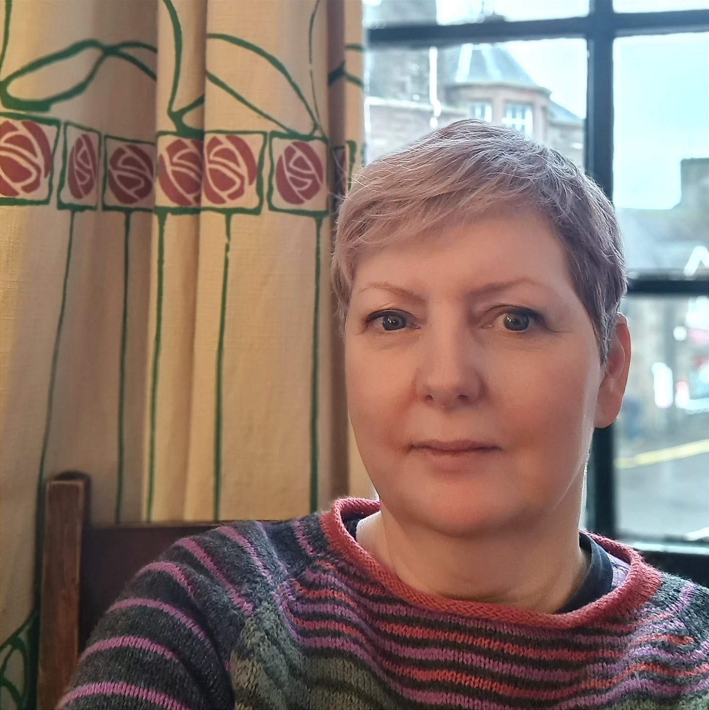 Photo of a woman in her 50s with short grey hair, seated at a window with cream coloured curtains with an Art Nouveau design featuring a stylised rose.
