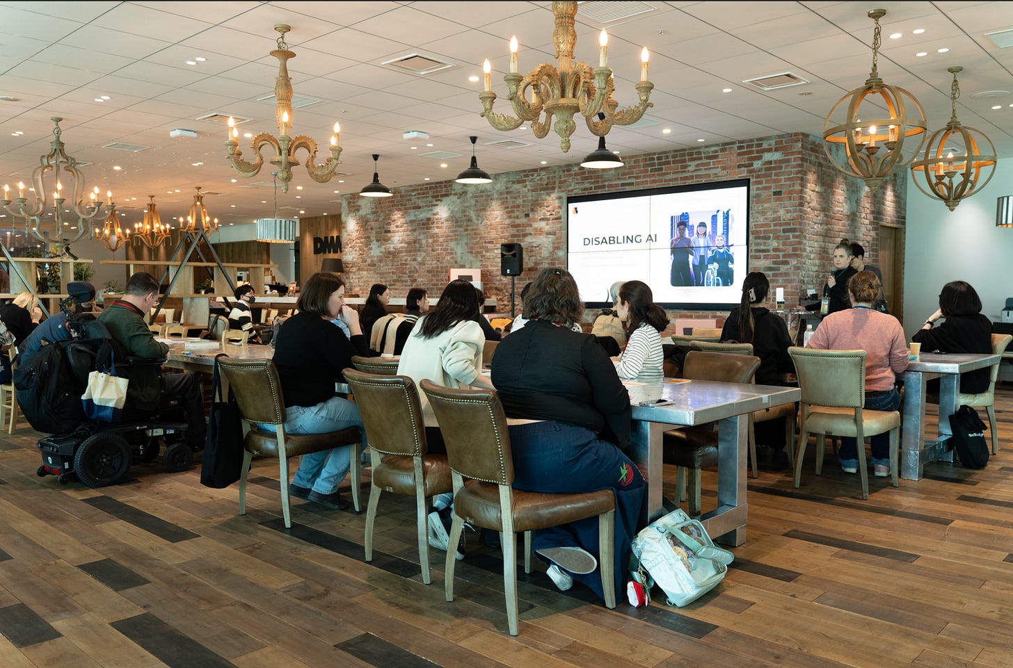    A diverse group of attendees sit at tables in a stylish, warmly lit event space, attentively watching a presentation titled "Disabling AI" projected on a large screen at the front of the room. The venue has an eclectic, upscale feel with exposed brick walls, mixed chandelier styles ranging from ornate gold candelabras to rustic sphere fixtures, and rich hardwood flooring. A speaker stands near the screen to the right. The atmosphere feels engaged and focused, suggesting a professional conference or panel discussion centered around disability and artificial intelligence.