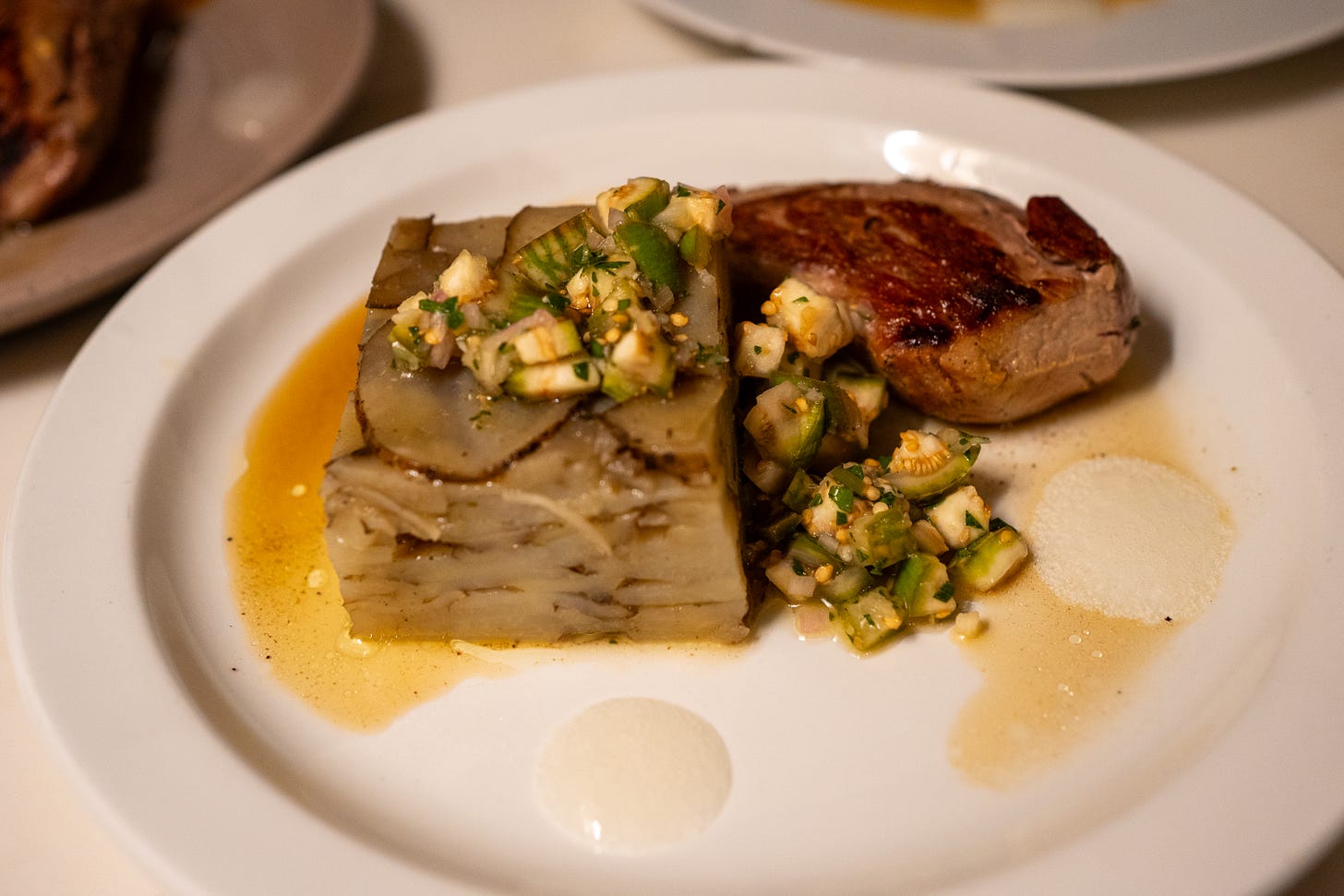 A white plate holding a portion of seared Berkshire pork tenderloin, a square layered russet potato terrine topped with a chunky green eggplant relish, all resting in a light amber jus.