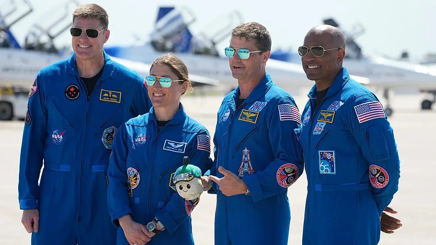  Gemini said Four astronauts in blue flight suits and sunglasses stand on a sunny tarmac in front of T-38 jet trainers. From left to right: Jeremy Hansen, Christina Koch, Reid Wiseman (pointing to a small plush doll), and Victor Glover. All are smiling.