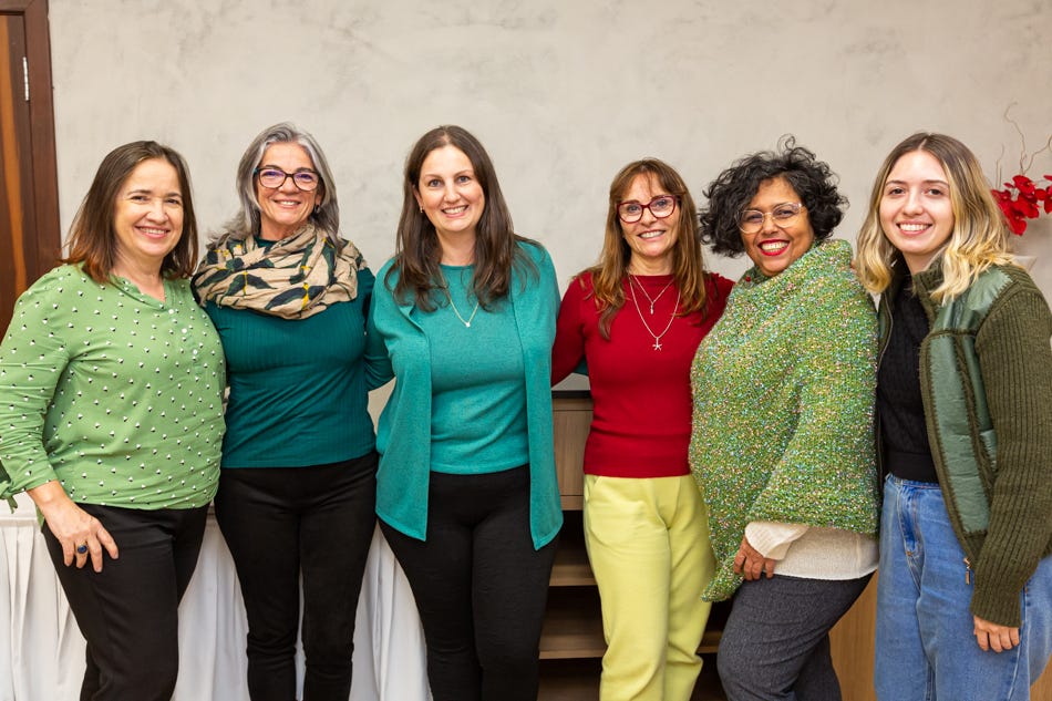 Foto de seis mulheres reunidas, sorrindo para a câmera. Elas estão em pé, posando em grupo. Ao fundo, há uma parede cinza, uma mesa coberta com toalha branca e uma flor vermelha no canto direito. Da esquerda para a direita: Dolores é uma mulher branca, com cabelos lisos castanhos na altura dos ombros. Ela veste uma blusa verde e calça preta. Fernanda é uma mulher branca, com cabelos grisalhos compridos, óculos e uma echarpe bege e verde. Ela veste blusa verde e calça preta. Mariana é uma mulher branca, com cabelos castanhos compridos. Ela usa um casaco verde, blusa verde e calça preta. Cristina é uma mulher branca, com cabelos castanho-claro compridos e óculos. Ela veste uma blusa vermelha e calça amarela. Silvia é uma mulher negra, com cabelos curtos e encaracolados pretos, óculos e um poncho verde. Ela usa uma blusa branca e calça cinza. Daniele é uma mulher branca, com cabelos loiros. Ela veste um casaco verde, blusa preta e calça jeans. (fim da descrição)