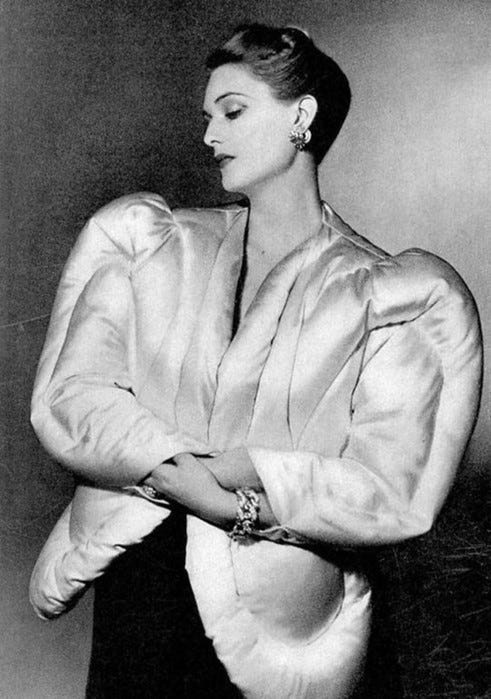 A black and white photo depicts a woman from the three-w\quarter perspective wearing a sculptural waist-length puffer coat in white silk.