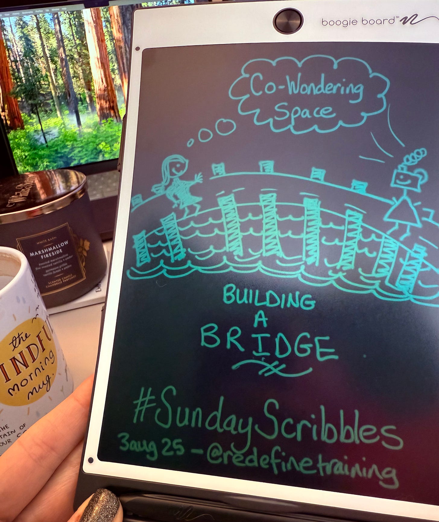 A close-up of Georgina Dean's hand holding a black Boogie Board. The screen displays her turquoise sketchnote for the '#SundayScribbles' creative prompt, showing a bridge connecting two figures with the theme 'Building A Bridge'. A cozy desk with a monitor and a 'Marshmallow Fireside' candle is in the background.