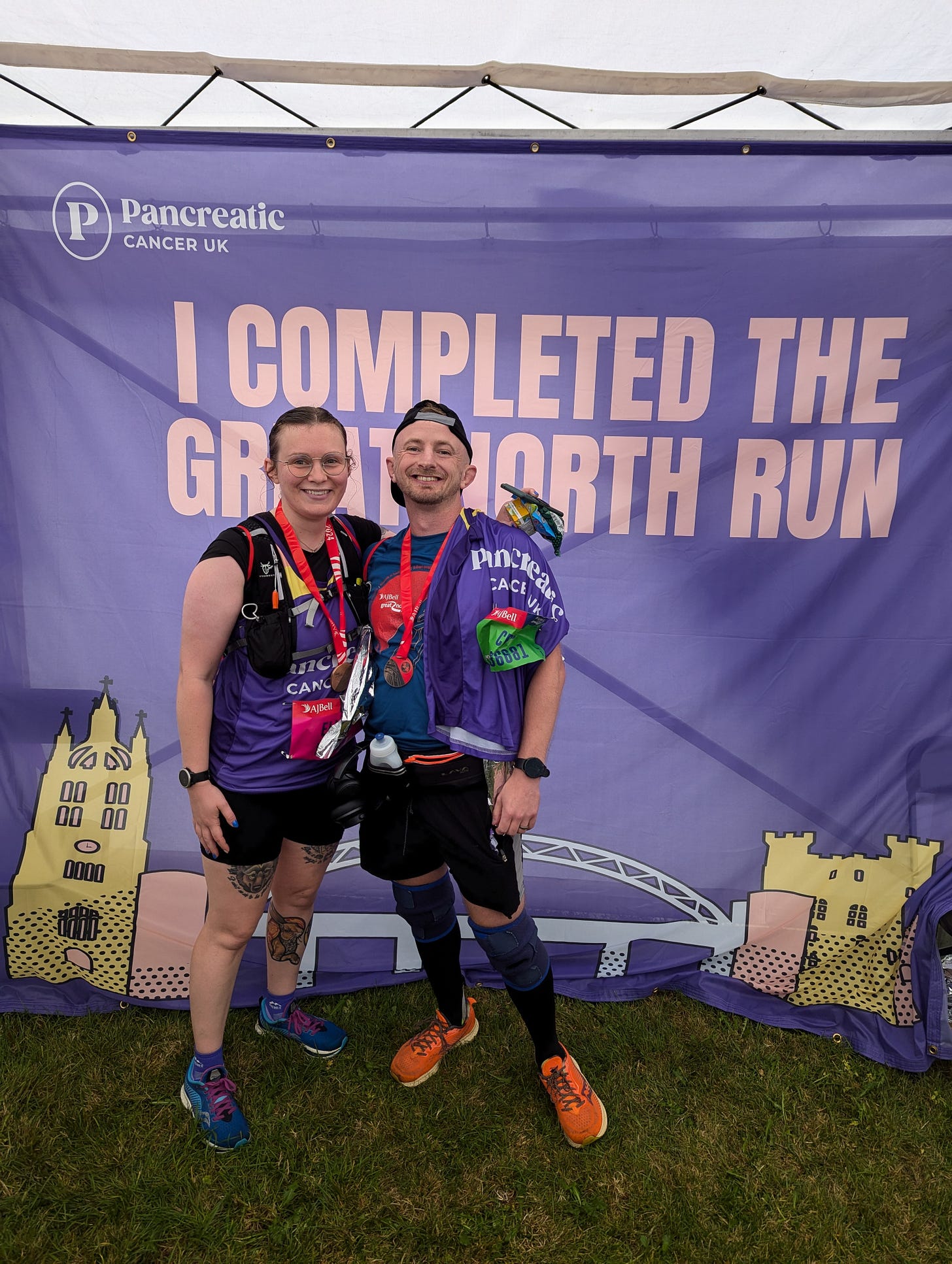Ellen and Craig standing in front of a purple sign that reads: I completed the Great North Run with the Pancreatic Cancer logo. Both are wearing medals and look very tired but happy.