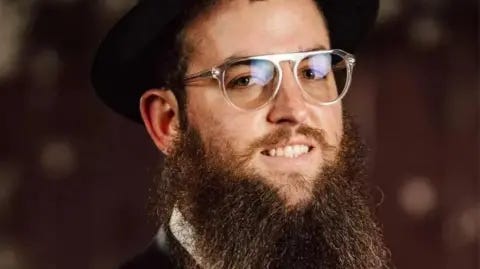 Chabad Zvi Kogan is seen in a portrait photograph. A bearded man, he is smiling at the camera and wearing glasses with clear plastic frames. There is a black hat, a fedora, on his head.