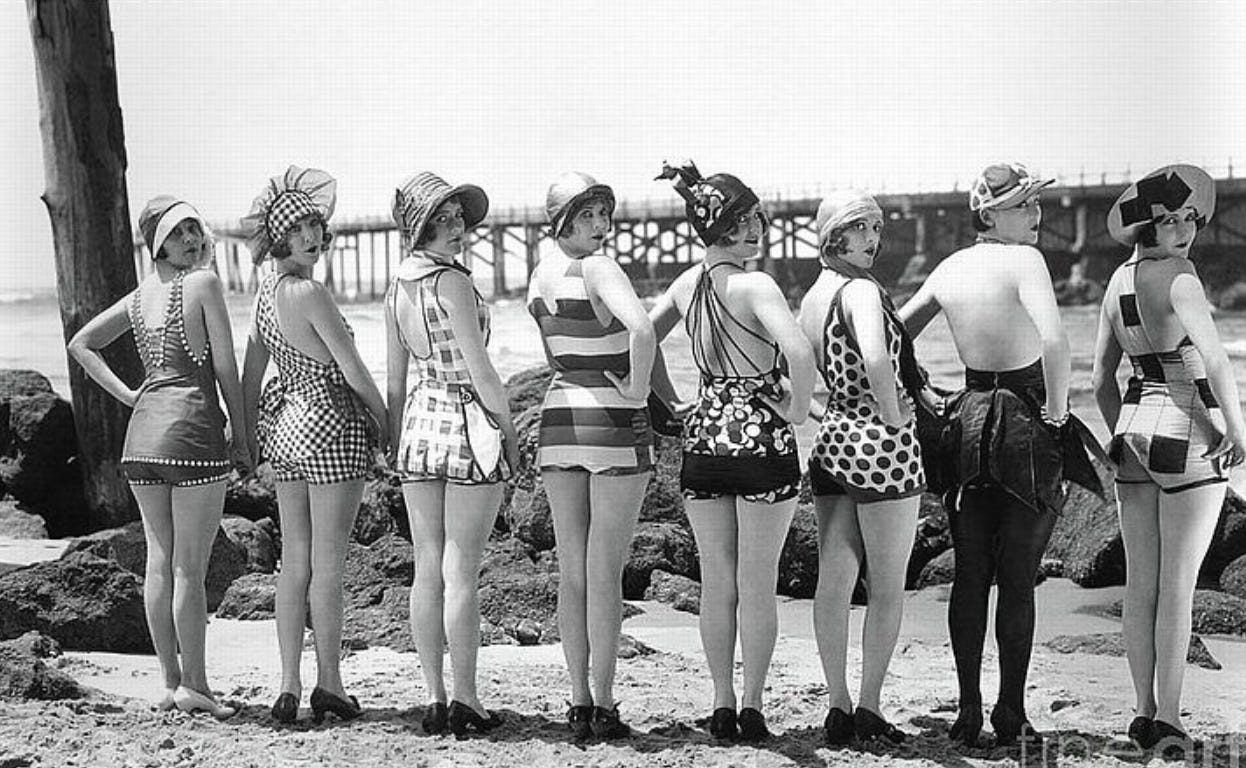 A vintage black-and-white photograph of nine women standing in a row on a beach, all turned to look over their shoulders toward the camera. They are dressed in a variety of patterned and striped 1920s-era swimsuits and swim caps, each with a distinct design. A wooden pier and rocky shoreline appear in the background. A vintage black-and-white photograph of nine women standing in a row on a beach, all turned to look over their shoulders toward the camera. They are dressed in a variety of patterned and striped 1920s-era swimsuits and swim caps, each with a distinct design. A wooden pier and rocky shoreline appear in the background.
