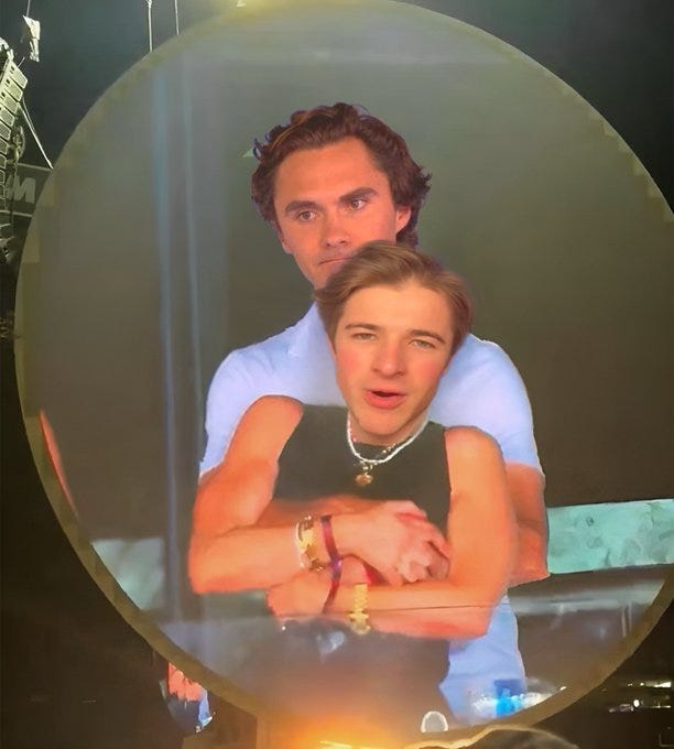 The image shows two men, one standing behind the other in a playful pose, displayed on a large oval screen, likely at a public event or concert. The man in front, wearing a sleeveless shirt and necklace, has his arms crossed, while the man behind, in a light shirt, has his arms around him. The setting appears lively with bright lights and a stage-like background. The post text, including the reply to Harry Sisson and the original comment about "beta MAGA losers" and hitting the gym, suggests a political or cultural critique, possibly mocking certain political supporters. No platform watermarks are visible. The image shows two men, one standing behind the other in a playful pose, displayed on a large oval screen, likely at a public event or concert. The man in front, wearing a sleeveless shirt and necklace, has his arms crossed, while the man behind, in a light shirt, has his arms around him. The setting appears lively with bright lights and a stage-like background. The post text, including the reply to Harry Sisson and the original comment about "beta MAGA losers" and hitting the gym, suggests a political or cultural critique, possibly mocking certain political supporters. No platform watermarks are visible.