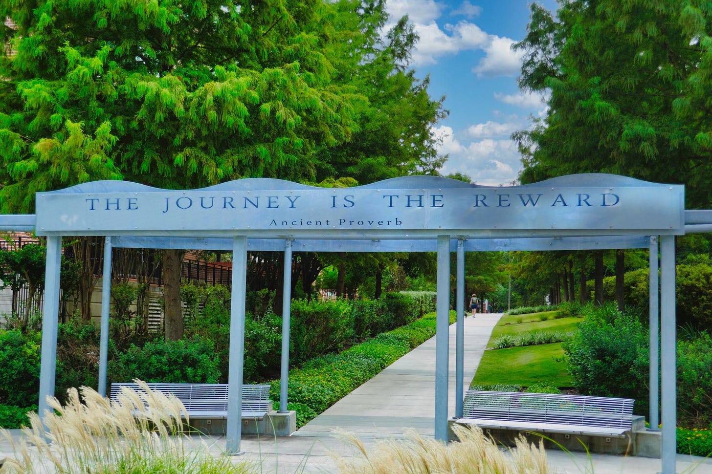 An outdoor structure with benches beneath the top attached with posts. On the roof a sign reads: “The Journey Is the Reward”