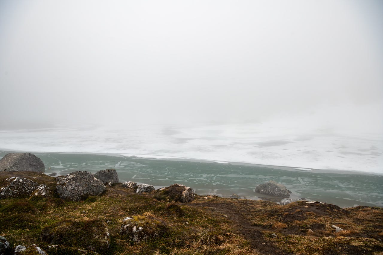 The shoreline of a mostly frozen lake seen from a low tundra bluff. A band of open water along the near edge shows pale green under a cracked, translucent ice sheet, which whitens with snow cover farther out before fog swallows the far shore entirely. Brown tundra and mossy boulders frame the near shore.