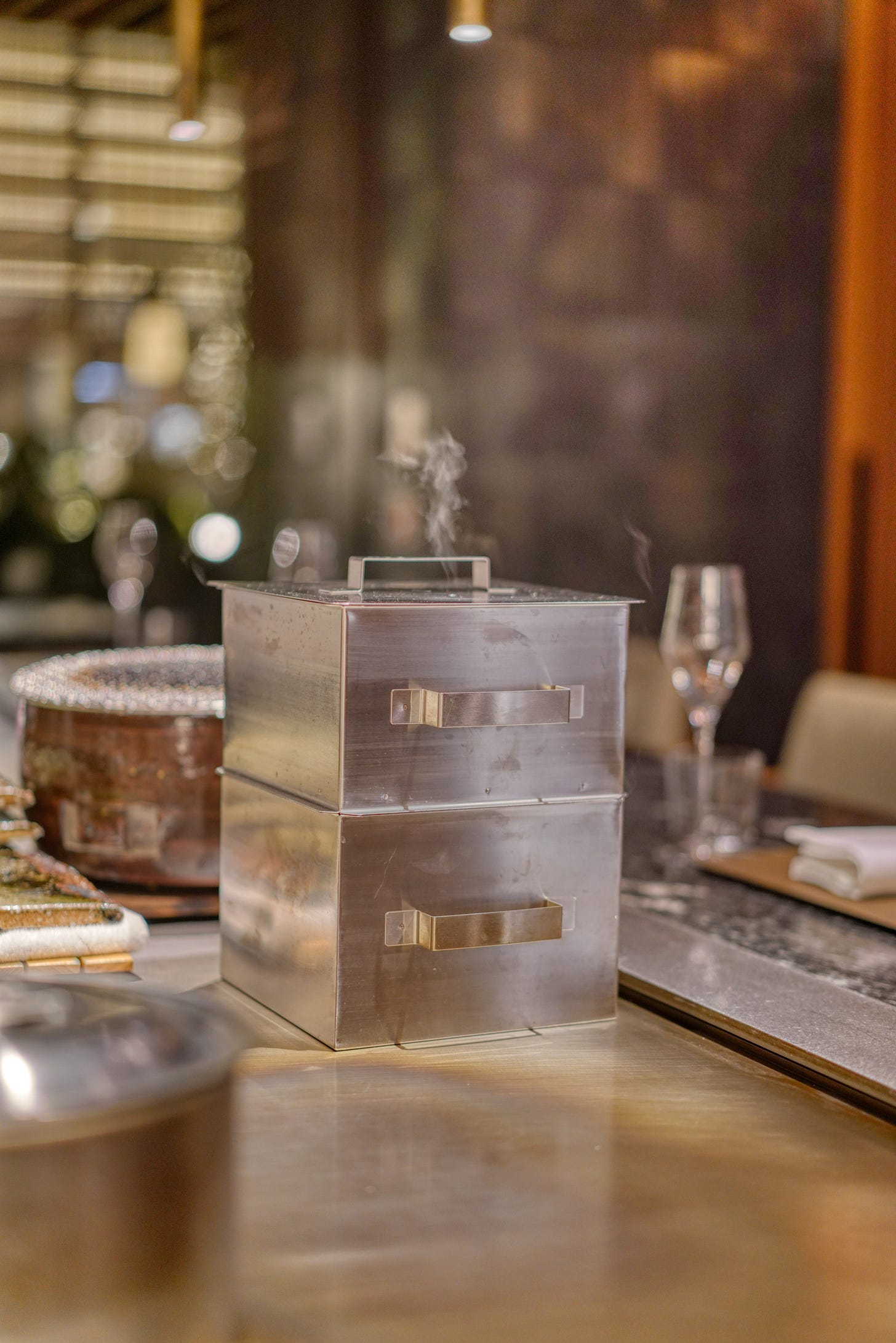 Stacked stainless-steel food steamers with steam rising from the top, sitting on a restaurant counter with blurred glassware and warm bokeh in the background. Stacked stainless-steel food steamers with steam rising from the top, sitting on a restaurant counter with blurred glassware and warm bokeh in the background.