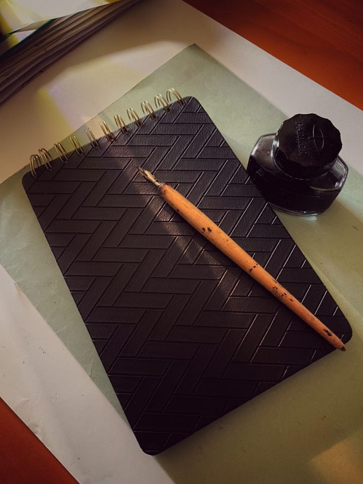 A split image displaying an artist's workspace. The left panel shows a black sketchbook with a herringbone texture, a wooden dip pen, and a Parker ink bottle sitting on a desk. The right panel is a close-up detail shot of the sketchbook's gold spiral binding, with the wooden pen slightly blurred in the background.