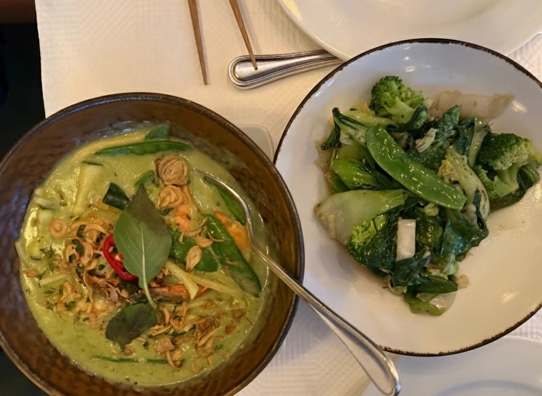 A warm bowl of green curry vegetables with basil and a side plate of sautéed greens on a restaurant table.