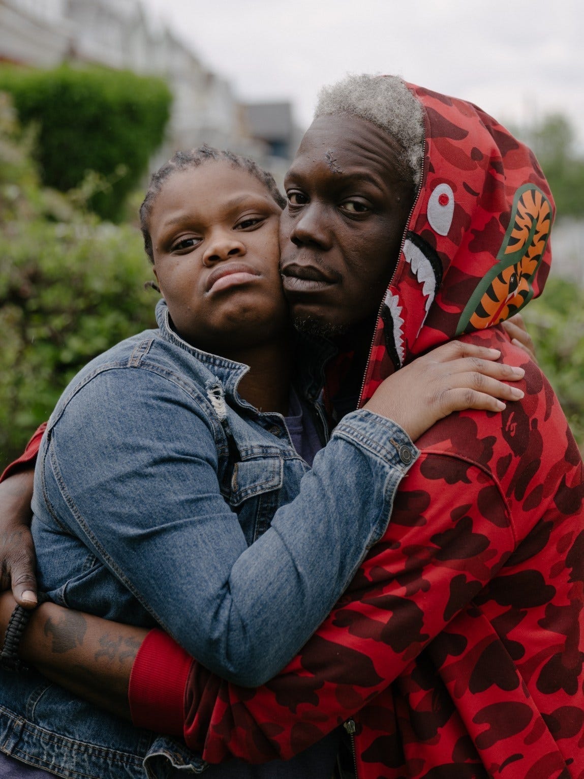 A daughter, wearing a jean jacket, and a father, wearing a brightly patterned red hoodie, pose holding each other and leaning their heads together. A daughter, wearing a jean jacket, and a father, wearing a brightly patterned red hoodie, pose holding each other and leaning their heads together.