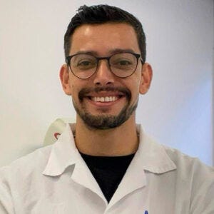 Homem de pele clara, cabelos curtos escuros e barba aparada. Usa óculos de armação escura e sorri para a câmera. Veste jaleco branco e camiseta preta por baixo. Homem de pele clara, cabelos curtos escuros e barba aparada. Usa óculos de armação escura e sorri para a câmera. Veste jaleco branco e camiseta preta por baixo.