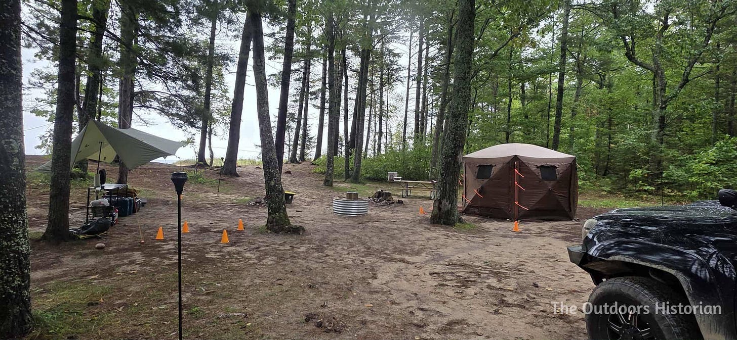 A view of a campsite with a pop-up Clam Tent, a fire pit, a tarp over a kitchen prep area, and tiki torches on a wooded bluff above Lake Superior. The front end of a Toyota Tacoma is also visible in the picture.