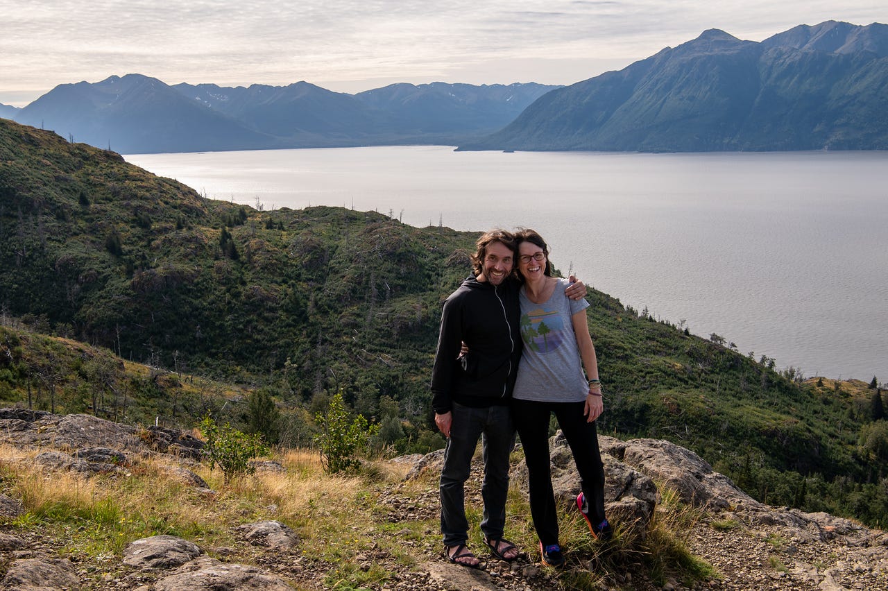 Two people stand arm in arm at an overlook above Turnagain Arm near McHugh Creek. They are smiling at the camera, one in a black jacket and sandals, the other in a gray T-shirt and sneakers. Behind them, steep green ridges drop down to the calm waters of Turnagain Arm, with tall mountains rising in the distance under a partly cloudy sky.