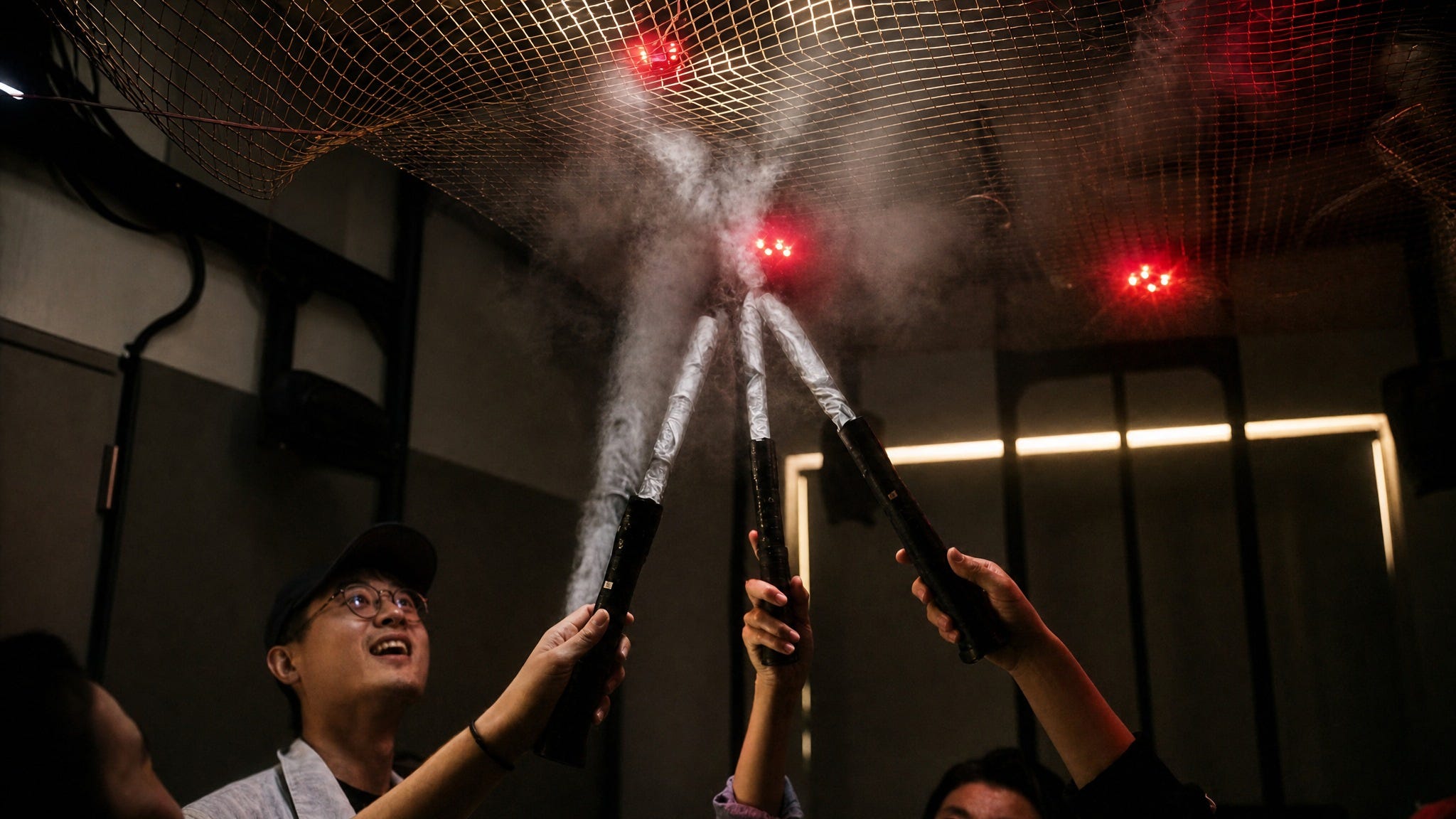 Three people under a suspended white net lit by red LED clusters, raising black flashlights whose beams streak across the mesh, one of them laughing Three people under a suspended white net lit by red LED clusters, raising black flashlights whose beams streak across the mesh, one of them laughing