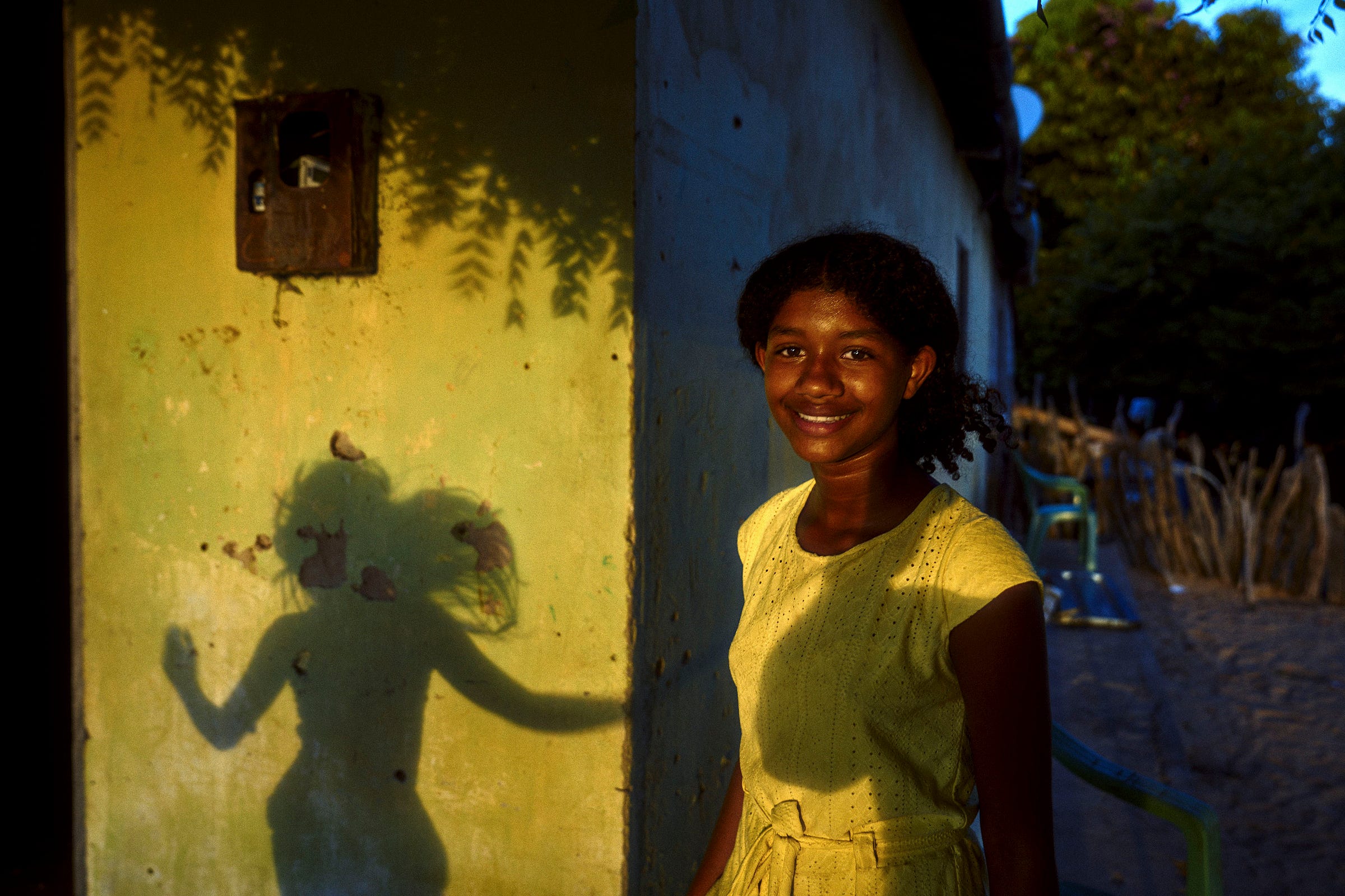 A smiling girl in a yellow dress stands beside a sunlit wall, casting a playful silhouette of herself with raised arms. The scene is bright with warm sunlight, and there are trees and a wooden fence in the background.
