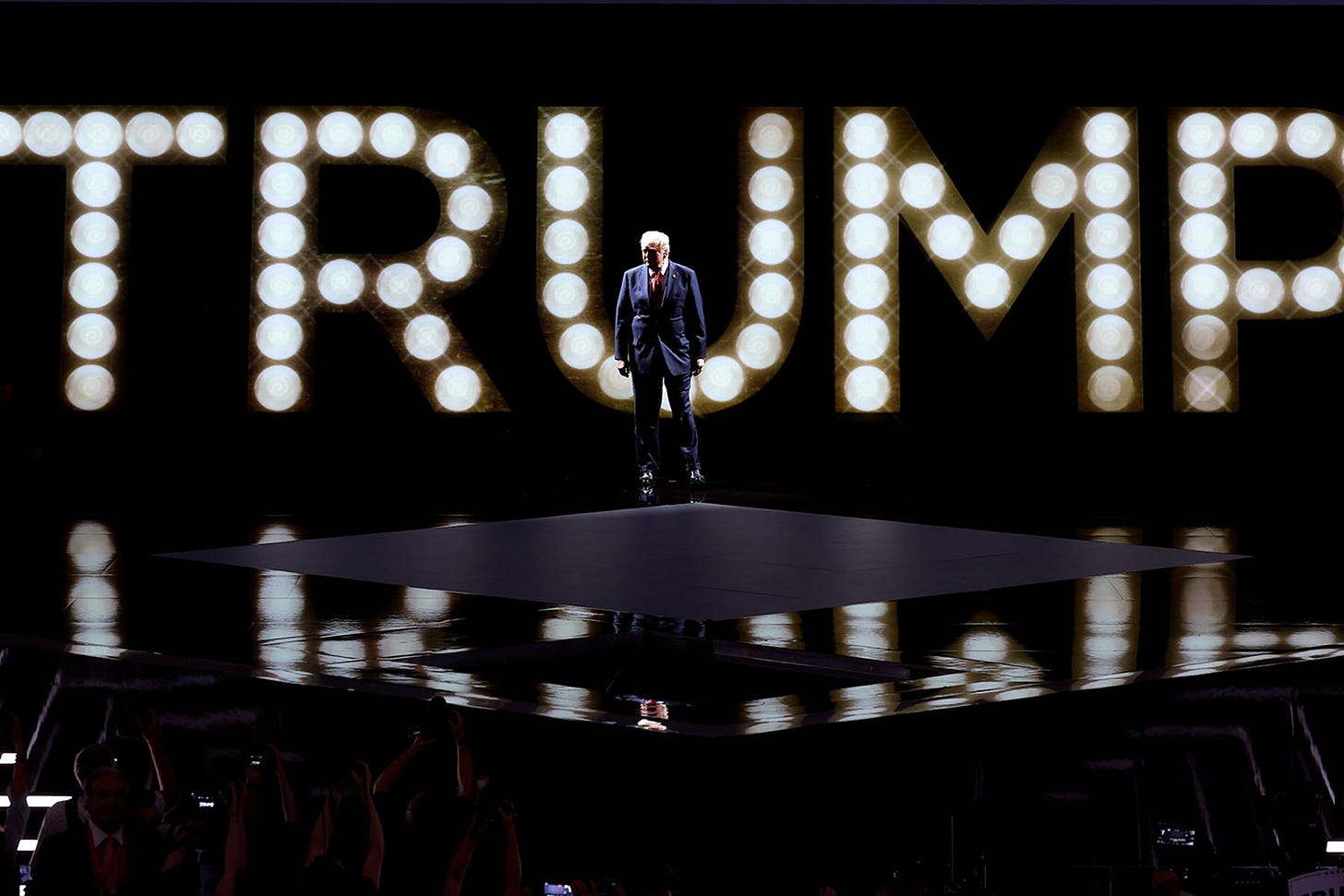 Trump stands on stage in front of lights spelling out "Trump"