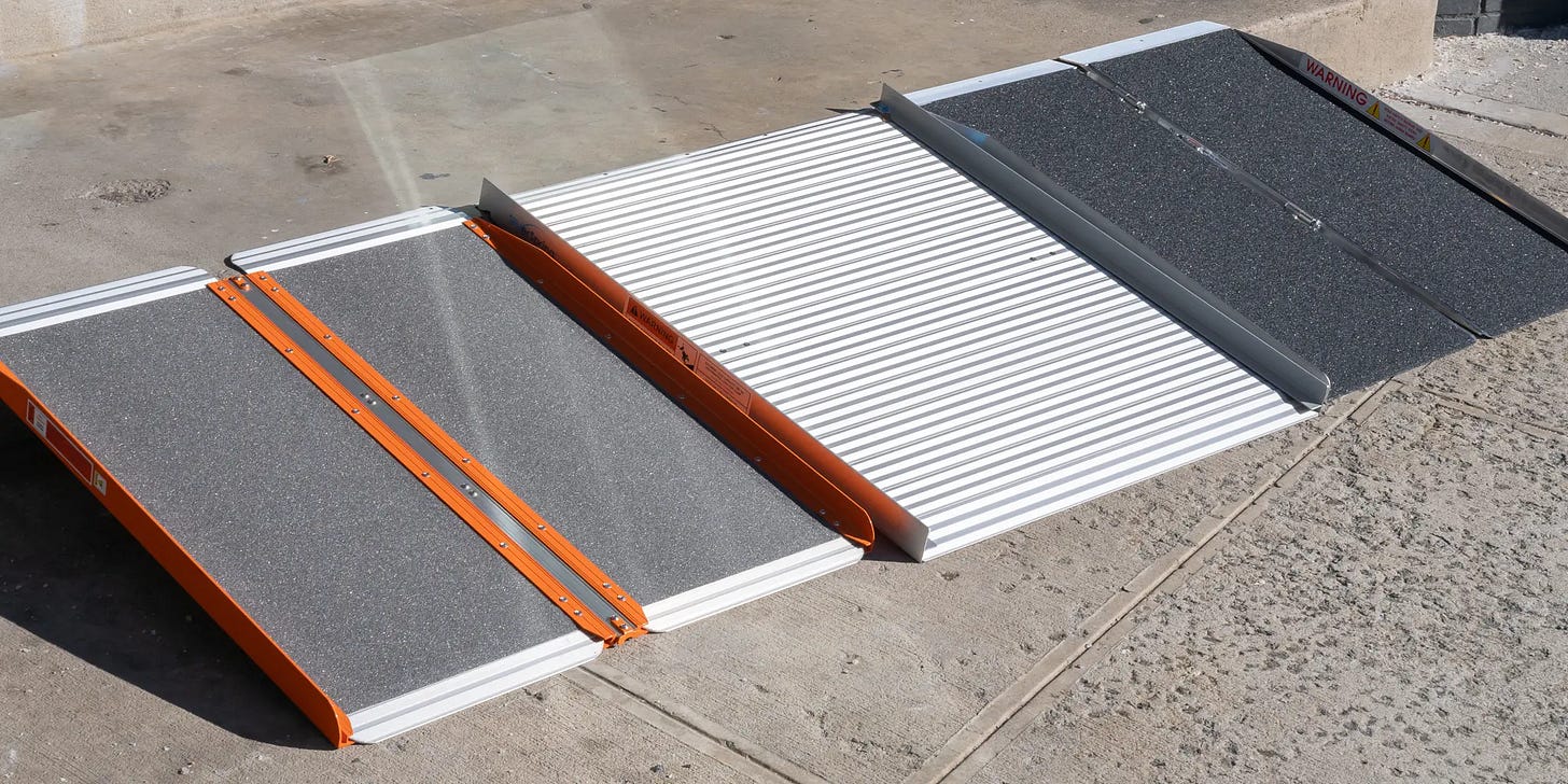 A portable metal accessibility ramp placed over a curb, made of three connected sections with textured, non-slip surfaces. The outer sections are dark gray with orange side rails, and the middle section is silver with ridges. A small warning label is visible on the raised end near the sidewalk.