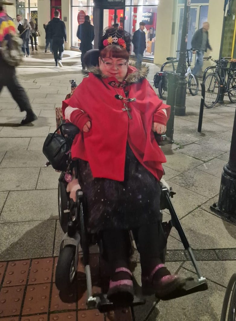 Penny sits in her wheelchair outside in Hastings in the darkening evening. She is wearing a bright red cape and has her hair worn high with a headband that is black with flowers and Halloween, Day of the Dead vibes, with a sugar skull in the centre. Besides wearing a black skirt she has red mittens on her hands for warmth. She is smiling gently. She wears glasses and her red hair shows in her short fringe.