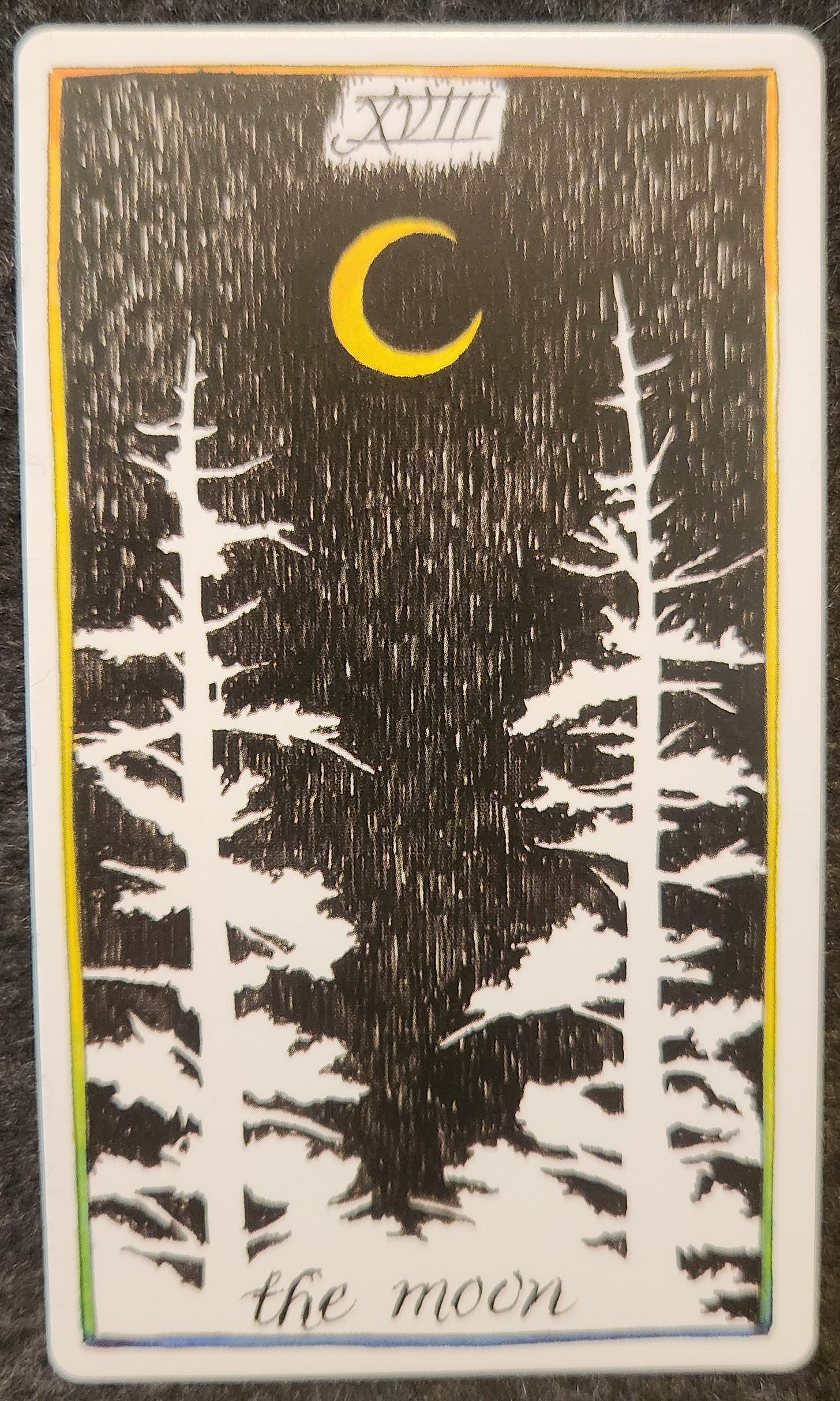 A tarot card labelled XVIII the moon A yellow crescent moon sits in the night sky between two tall pine trees. 