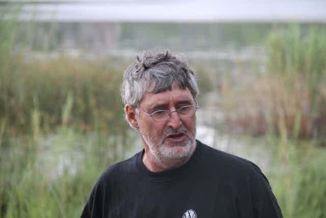 A man with greying hair and glasses speaks as his photo is taken against a backdrop of nature. A man with greying hair and glasses speaks as his photo is taken against a backdrop of nature.
