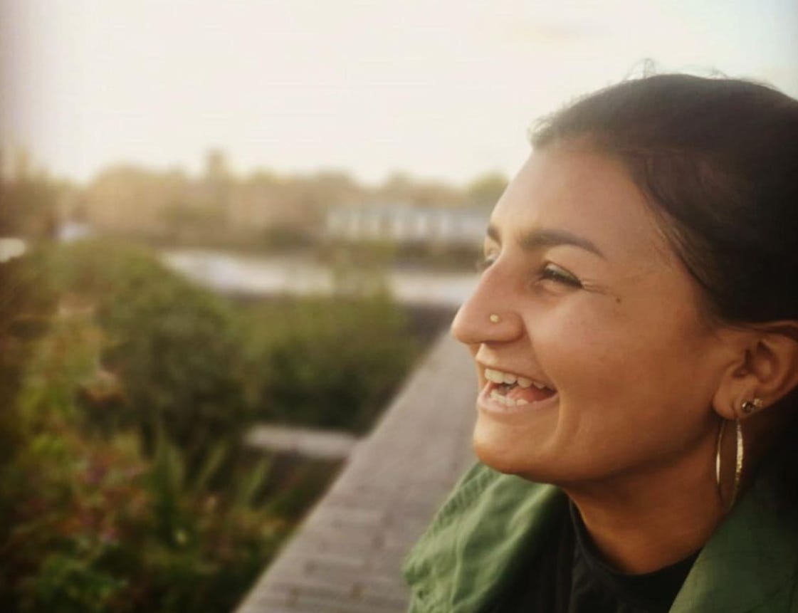 A young woman with big hoop earrings is looking out over a rooftop, smiling and laughing. It's a nice day and the blue sky and trees are blurred in the background.