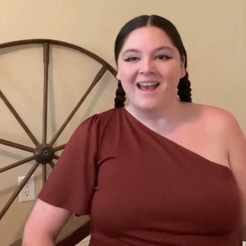 A repeating gif of a white woman with two braids, bold car eye makeup, and a brown t-shirt pulled off one shoulder reading from cue cards and saying Hi gay! Behind her is an antique-looking carriage wheel. A repeating gif of a white woman with two braids, bold car eye makeup, and a brown t-shirt pulled off one shoulder reading from cue cards and saying Hi gay! Behind her is an antique-looking carriage wheel.