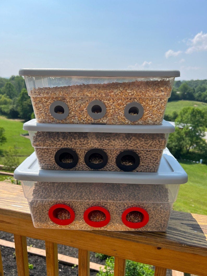 May include: Three clear plastic containers stacked on top of each other. Each container has three holes cut out of the side. The top container is filled with a light brown grain, the middle container is filled with brown pellets, and the bottom container is filled with a mixture of brown pellets and a light brown grain. The holes in the top container are gray, the holes in the middle container are black, and the holes in the bottom container are red.